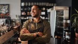 Young happy afro-american small coffee shop owner standing behind counter wearing apron with crossed arms.